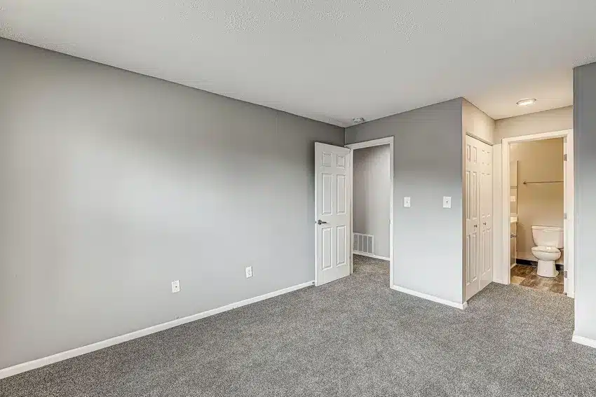 Empty bedroom with gray carpet, light gray walls, an open door, and view into a bathroom with a visible toilet and closet doors.