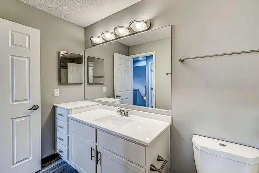 Modern bathroom with white vanity, large mirror, wall-mounted lights, toilet, and gray walls. Door and hallway are reflected in the mirror.