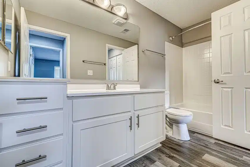 Modern bathroom with white cabinets, large mirror, single sink, toilet, and a bathtub with a showerhead. Door is open, and flooring is wood-patterned vinyl.