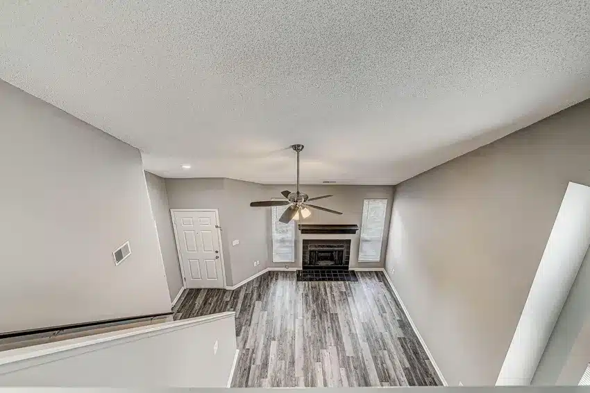 A view from above of a living room with gray wood flooring, a ceiling fan, a fireplace, two tall windows, and a white entry door.