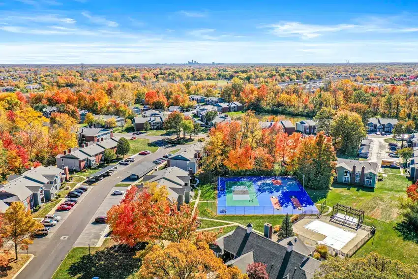Aerial view of a suburban neighborhood with autumn foliage, a blue tennis or pickleball court, residential houses, and distant city skyline under a clear blue sky.