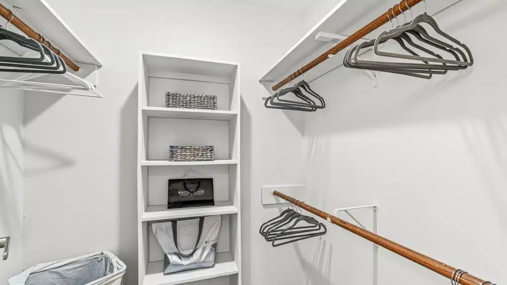 A walk-in closet with empty shelves, a few baskets, bags, and several hangers on wooden rods against white walls.