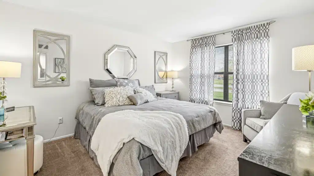 A neatly made bed with gray bedding sits between two nightstands and lamps in a bright, carpeted bedroom with patterned curtains and decorative mirrors.