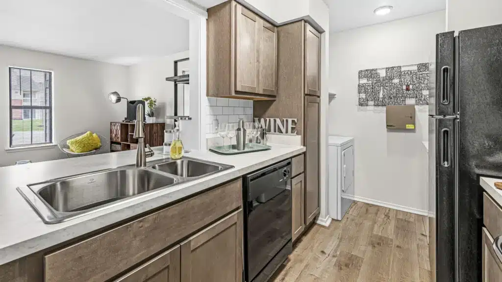 Modern kitchen with wood cabinets, double sink, dishwasher, and black appliances; view into a living area and laundry machines visible in the background.