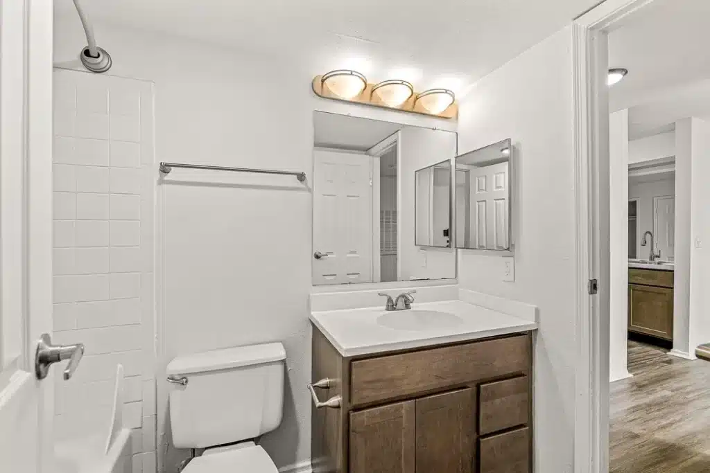 A bathroom with a wooden vanity, white countertop, wall-mounted mirror, toilet, towel rack, and a shower-tub combination with white tile.
