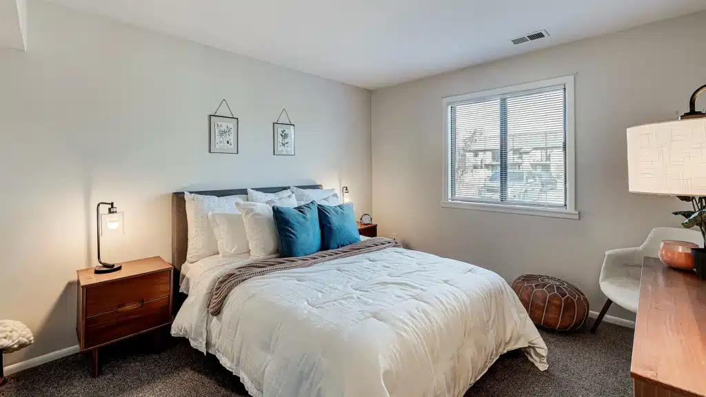 A neatly made bed with blue and white pillows is centered in a simple bedroom with two nightstands, a window with blinds, and minimal wall decor.