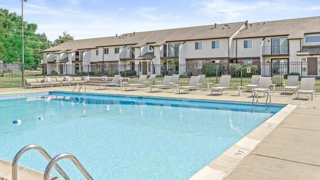 Outdoor swimming pool with lounge chairs, metal handrails, and a fenced area in front of a row of two-story apartment buildings under a clear sky.
