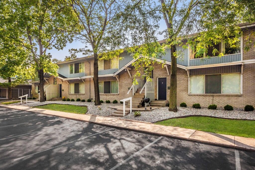 Two-story brick apartment building with stairs leading to each unit, surrounded by trees and landscaping, and facing a parking lot.