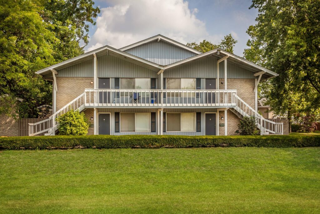 A two-story apartment building with exterior staircases, a white railing, and a well-maintained lawn in front, surrounded by trees and shrubs.