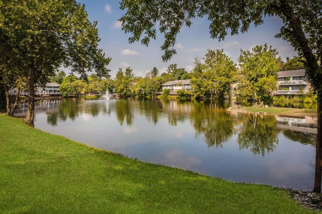 A calm pond with a fountain, surrounded by green grass, trees, and white residential buildings under a partly cloudy sky.