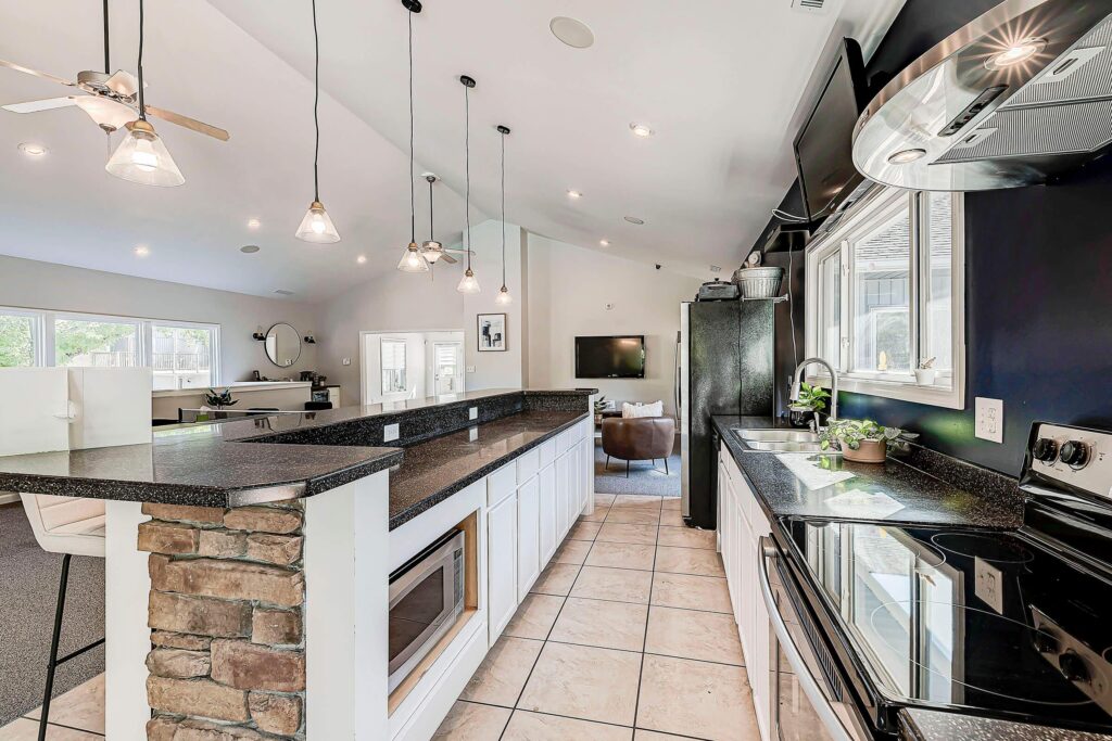 Modern kitchen with black countertops, white cabinets, a stone island, hanging pendant lights, tiled floor, and an adjacent living area with windows and a TV.