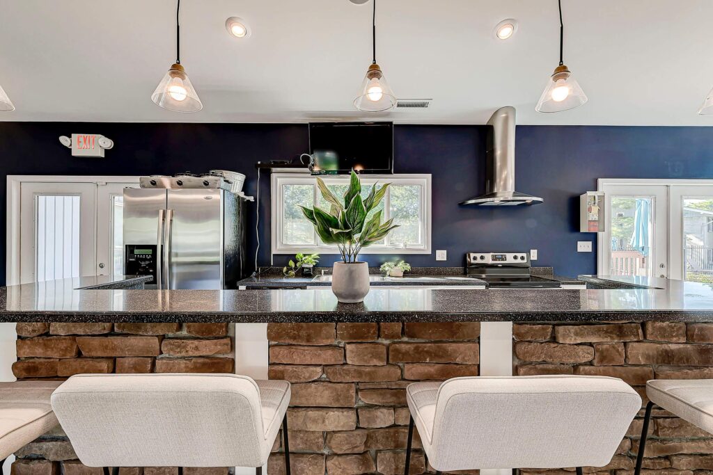 Modern kitchen with stone island, granite countertop, white bar stools, stainless steel appliances, and potted plant centerpiece; pendant lights hang from the ceiling.