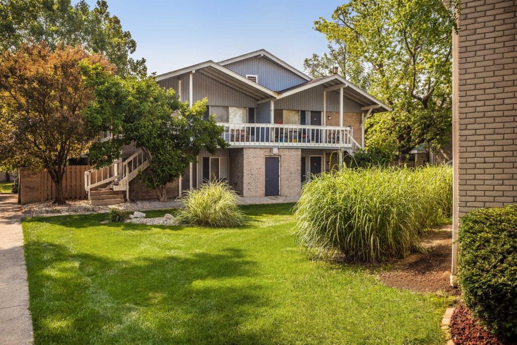 A two-story apartment building with a balcony and exterior staircase, surrounded by green grass, trees, and landscaped bushes on a sunny day.