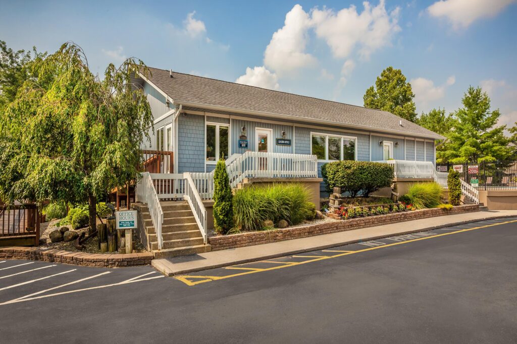 A single-story light blue building with a porch, stairs, and landscaping, located next to a parking lot with marked spaces under a partly cloudy sky.