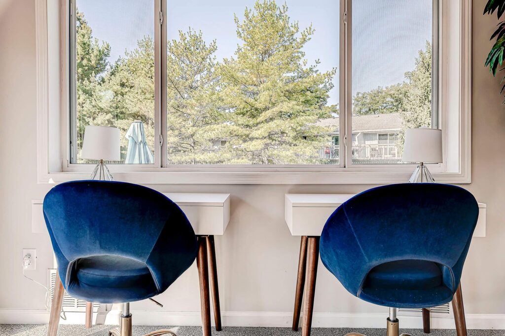 Two blue chairs face two white desks positioned under a large window with a view of trees and houses outside. Each desk has a small lamp on it.