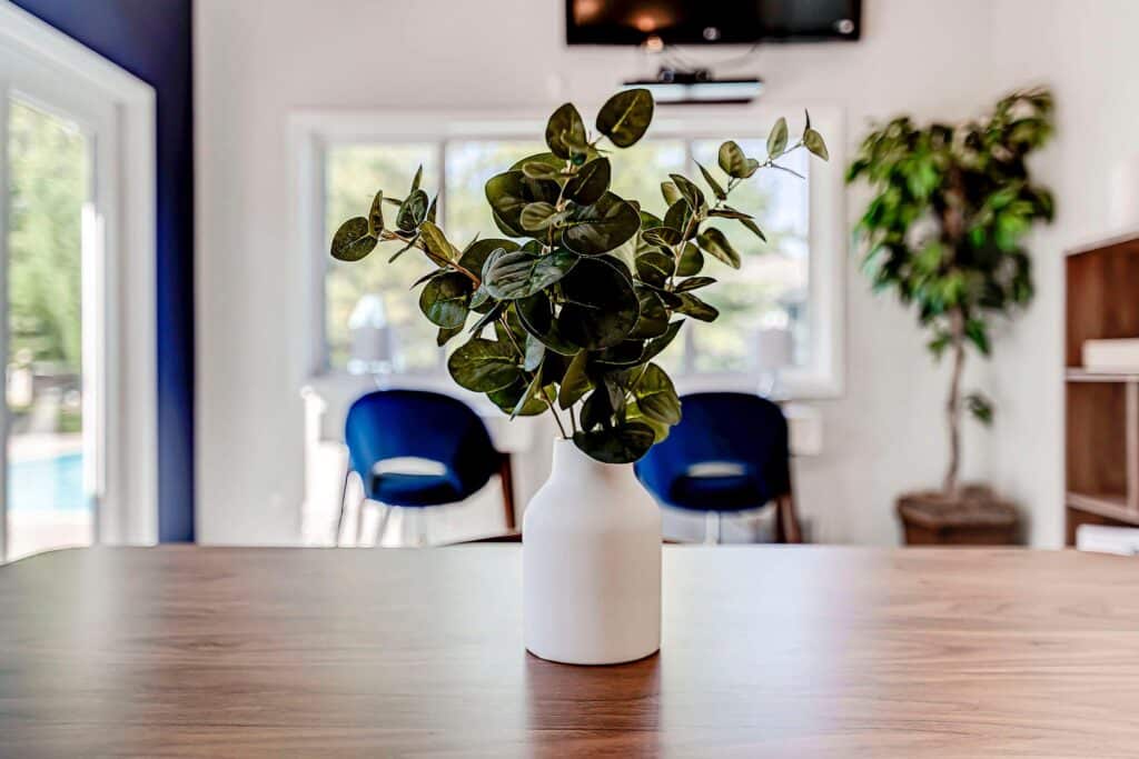 A white vase with green leafy branches sits on a wooden table in a modern, well-lit room with blue chairs and a potted plant in the background.