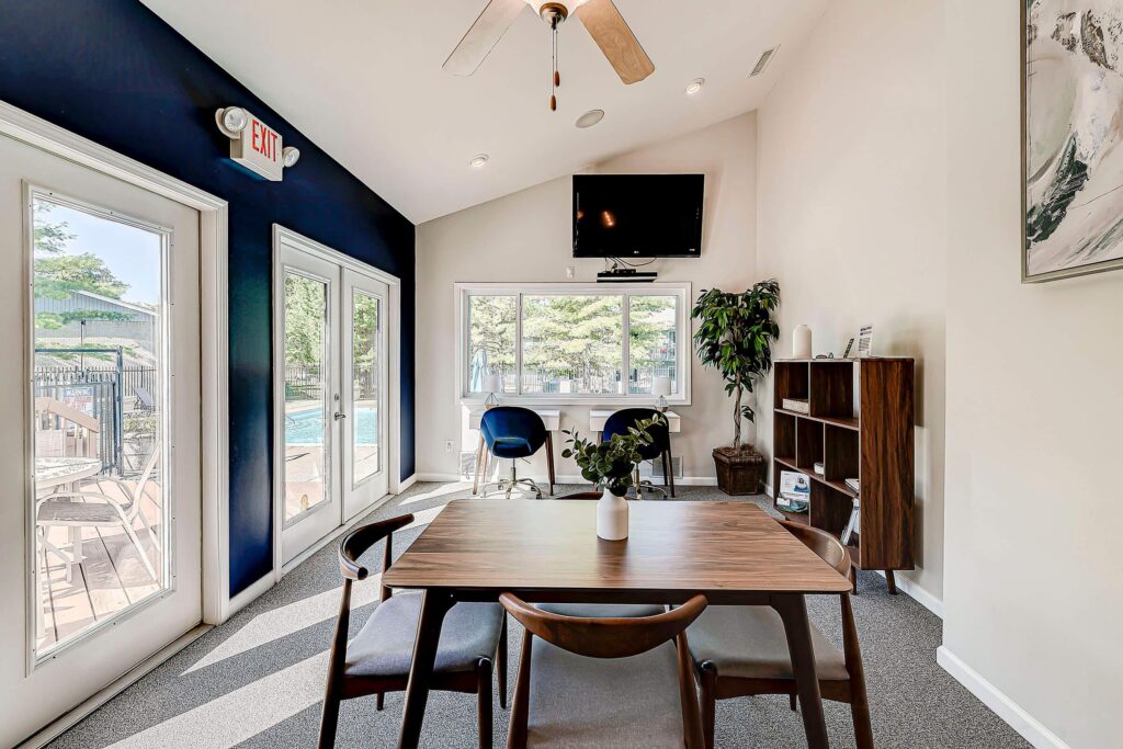 A modern meeting room with a wooden table, four chairs, a wall-mounted TV, bookshelves, large windows, and doors leading to an outdoor pool area.