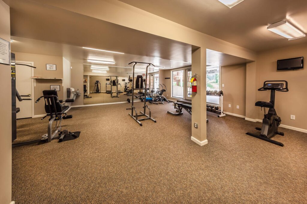 A small gym with exercise machines, carpeted floor, large mirrors on one wall, and a mounted TV in the corner.