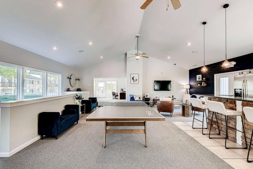 Spacious recreation room with a ping pong table, bar seating, lounge chairs, wall-mounted TV, ceiling fans, and large windows letting in natural light.