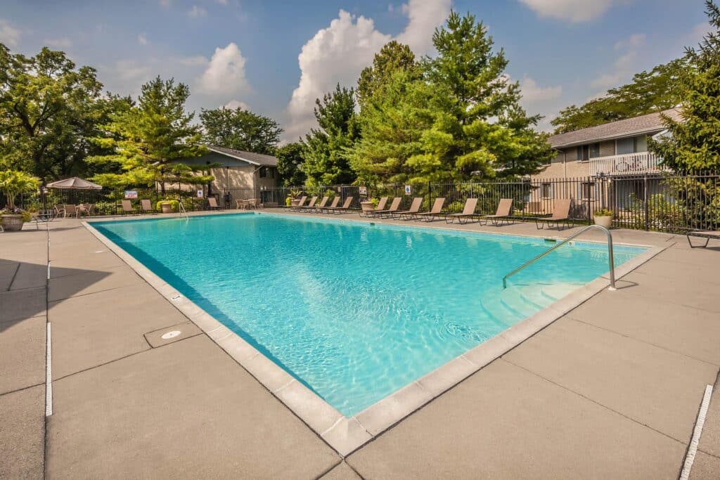Outdoor swimming pool with clear water, surrounded by lounge chairs and trees, located in an apartment complex under a partly cloudy sky.