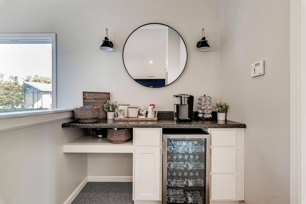 A small coffee bar setup with a round mirror, black countertop, mini fridge, coffee maker, mugs, baskets, and two wall-mounted lights against a light gray wall.