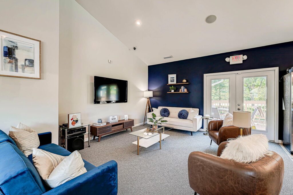 Modern living room with blue accent wall, two sofas, an armchair, a wall-mounted TV, coffee tables, and large windows leading to an outdoor deck.