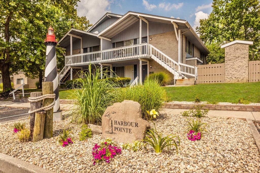 Two-story apartment building with balconies, landscaped garden, lighthouse decoration, and a stone sign reading "Harbour Pointe" at the entrance.