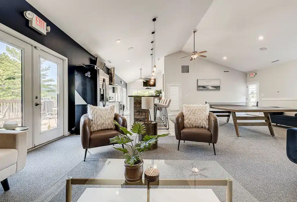 Modern open-concept living space with two armchairs, a glass coffee table, kitchen island, and a foosball table; large windows and ceiling fans provide natural light and ventilation.