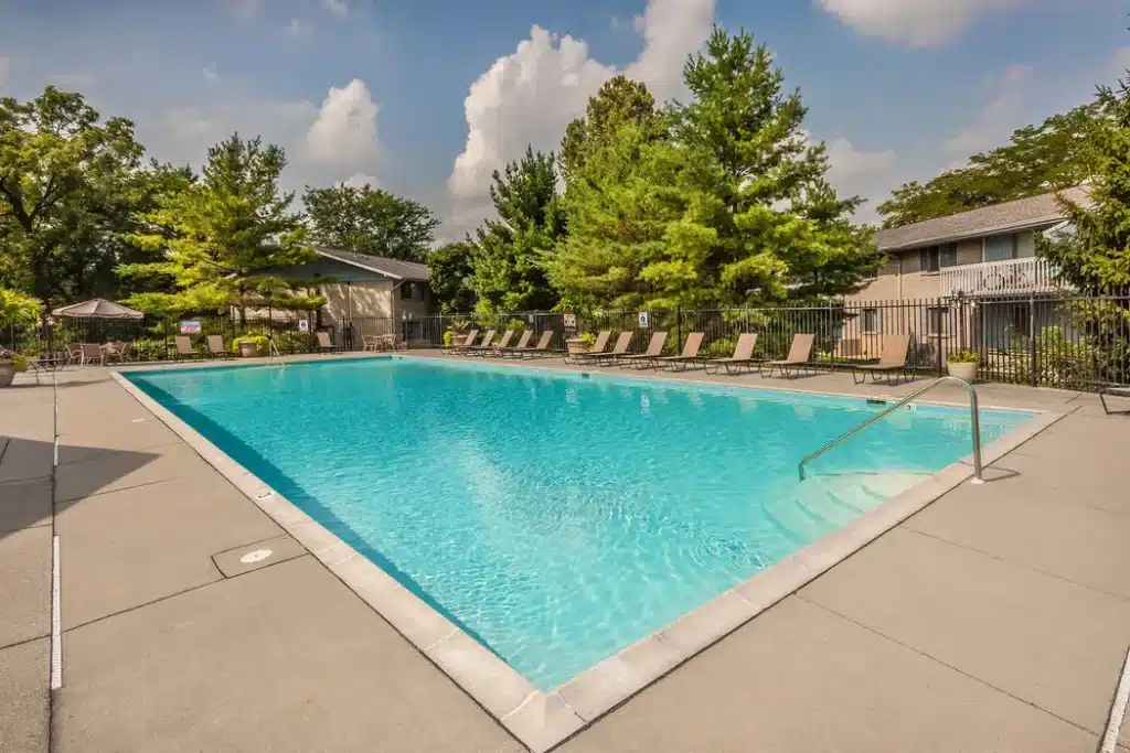 Outdoor swimming pool surrounded by lounge chairs, trees, and apartment buildings under a partly cloudy sky.