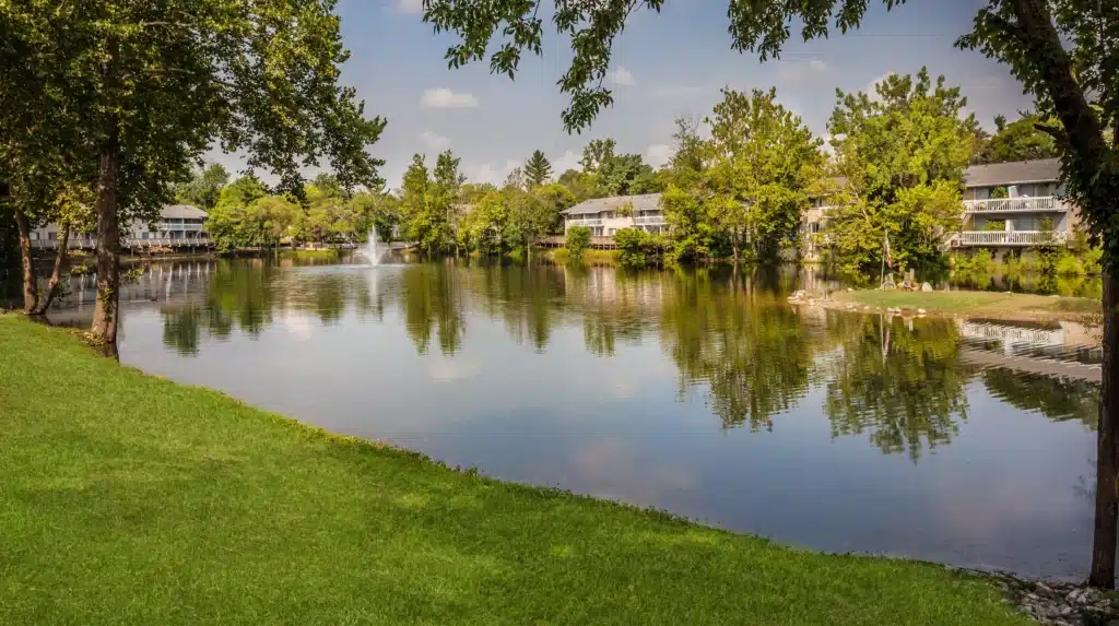 A calm lake bordered by green grass and trees, with apartment buildings in the background and a water fountain in the center.