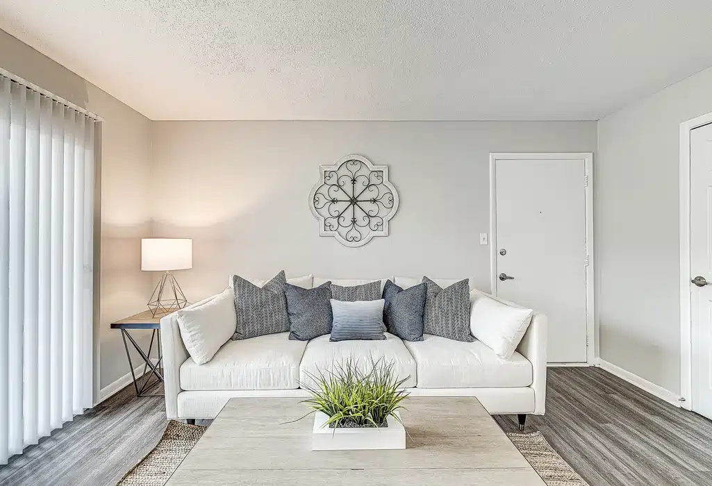 A modern living room with a white sofa, blue and gray pillows, a side table with a lamp, a wall decoration, and a coffee table with a plant. Vertical blinds cover a window.