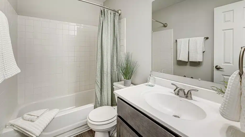 Modern bathroom with a bathtub and shower, green curtain, white towels, a sink with a silver faucet, a potted plant, and a mirror above the vanity.