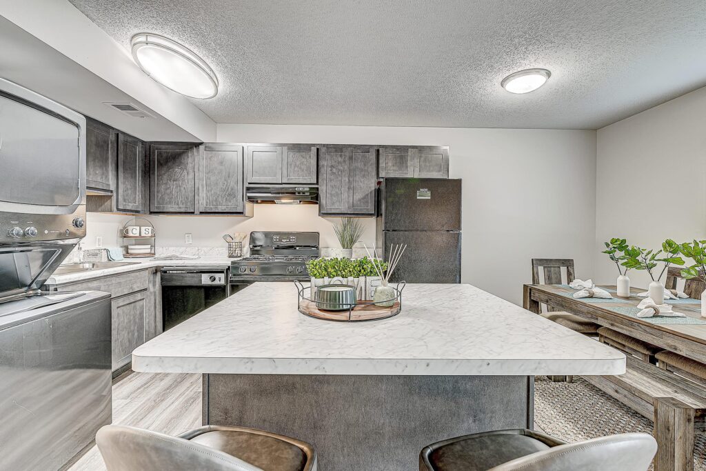 Modern kitchen with dark cabinets, white marble island, black appliances, and a dining table set with plates and green plants. Bright lighting and neutral decor throughout.