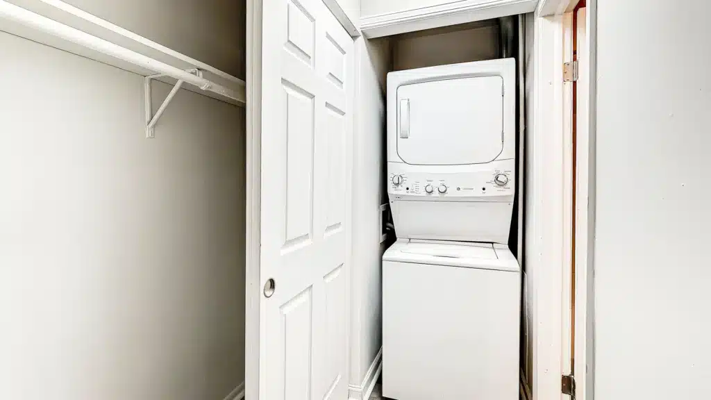 A white stacked washer and dryer unit is positioned in a small laundry closet with sliding doors, next to an empty closet with a white hanging rod.