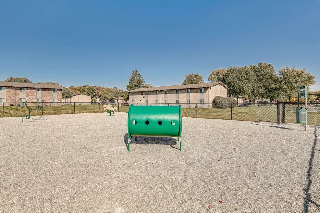 Fenced outdoor area with gravel ground, green agility equipment, and apartment buildings in the background under a clear sky.