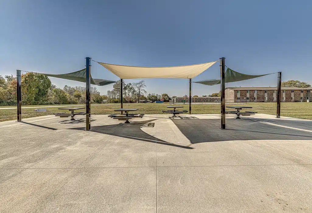 Outdoor picnic area with several tables under a rectangular shade canopy, surrounded by open grassy field and distant buildings under a clear sky.