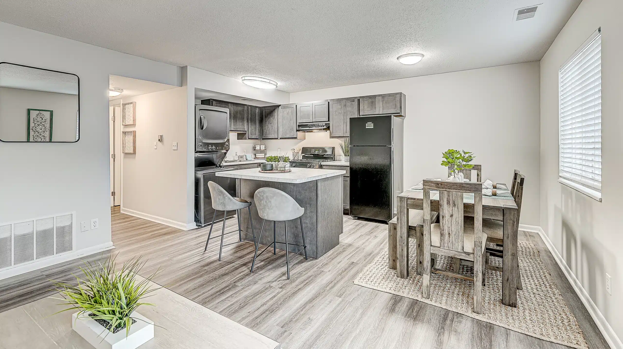 Modern kitchen and dining area with wood-style flooring, gray cabinets, black appliances, a dining table set, and two barstools at a small island under bright ceiling lights.