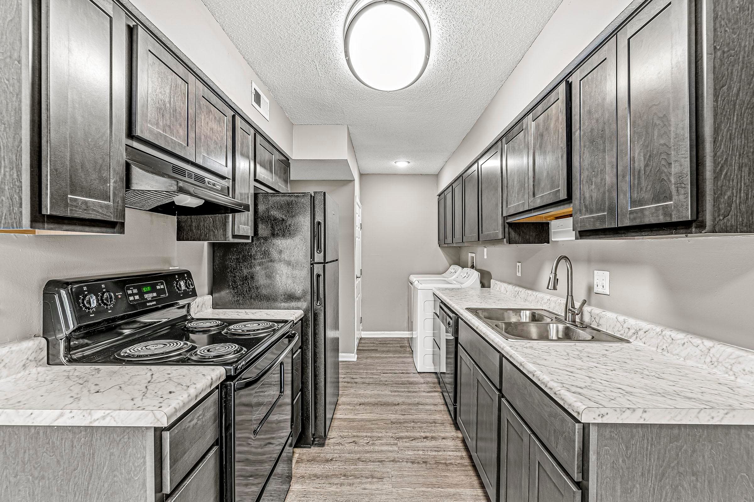 Galley kitchen with dark cabinets, marble-look countertops, black appliances, and stacked washer and dryer at the far end.