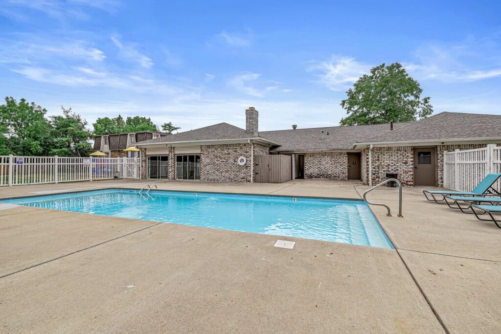 Rectangular outdoor swimming pool with metal handrails, surrounded by concrete deck, lounge chairs, and a brick building in the background under a blue sky.