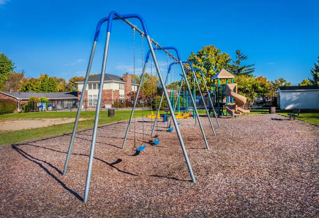 Empty playground with blue swings, wood chip ground, and a slide structure in the background, surrounded by green grass and trees under a clear blue sky.