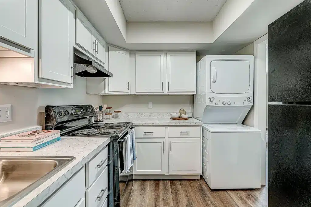Modern kitchen with white cabinets, black appliances, marble countertops, a stacked washer and dryer, and wood flooring.