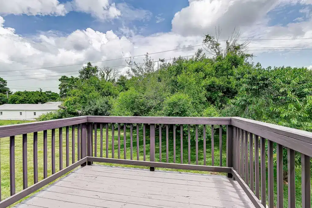 A wooden balcony with simple railing overlooks a grassy yard and dense green trees under a partly cloudy sky.
