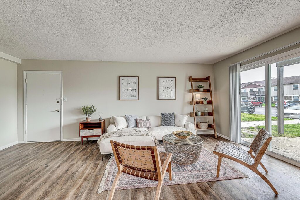 Modern living room with light-colored sofa, two woven chairs, a round coffee table, wood shelves, and large windows showing outdoor view and parked cars.