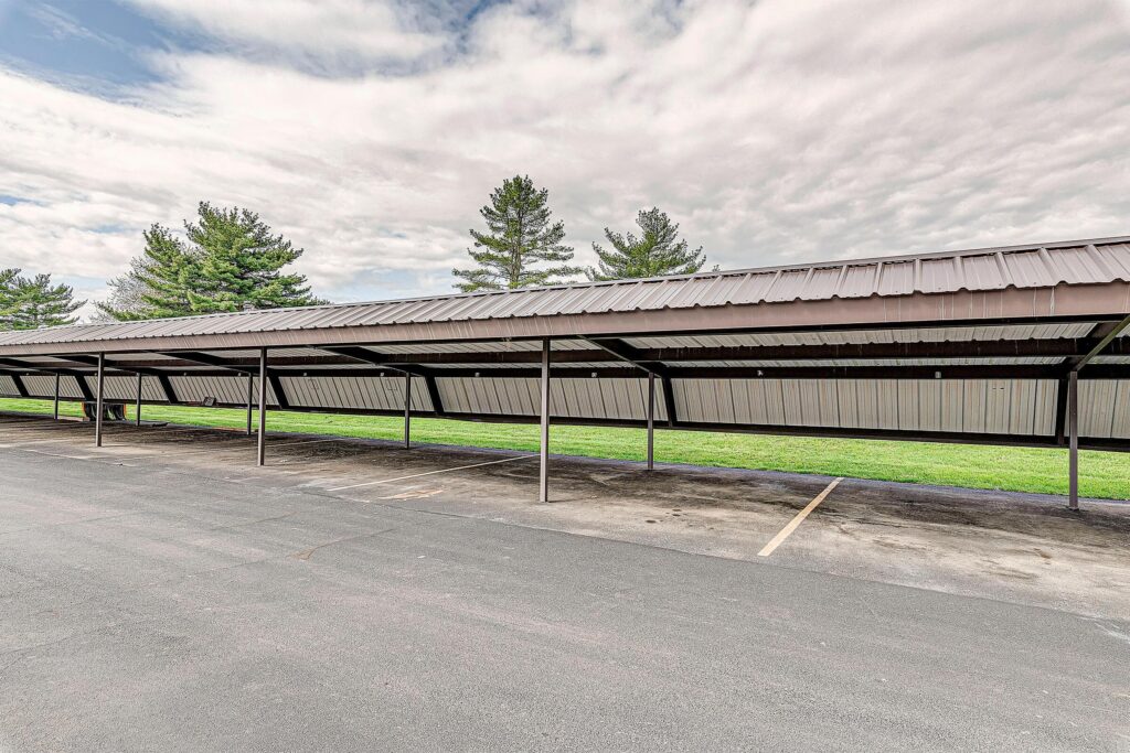 A covered parking area with metal roofing, empty parking spaces, and trees in the background under a cloudy sky.