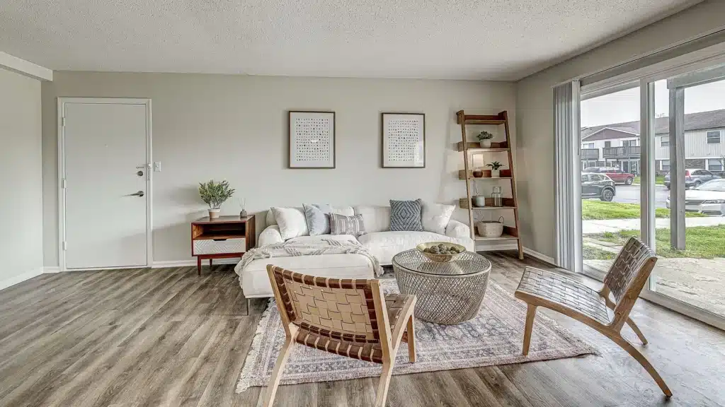 A modern living room with a white sofa, two woven chairs, a round coffee table, a side table, a ladder shelf, and large windows overlooking a street.