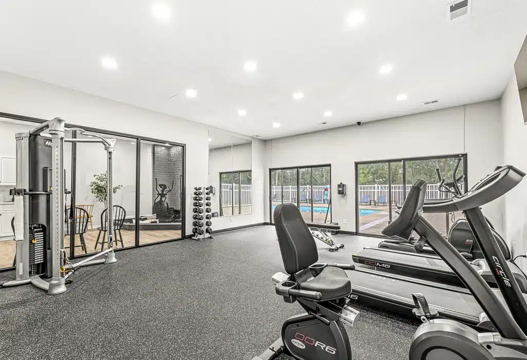 Modern gym with exercise bikes, treadmills, weight machine, and free weights; large windows overlook a pool area, and a mirrored wall reflects the equipment.