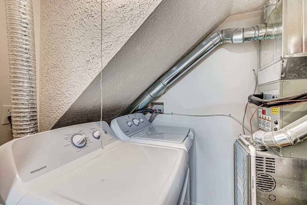 A washing machine and dryer are placed under a sloped ceiling next to a furnace with exposed air ducts.