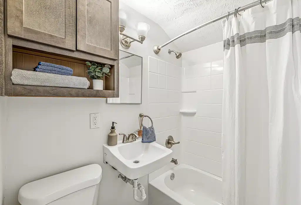 Small bathroom with a white sink, toilet, bathtub with a shower curtain, wall mirror, wooden cabinet, and neatly folded towels on a shelf.