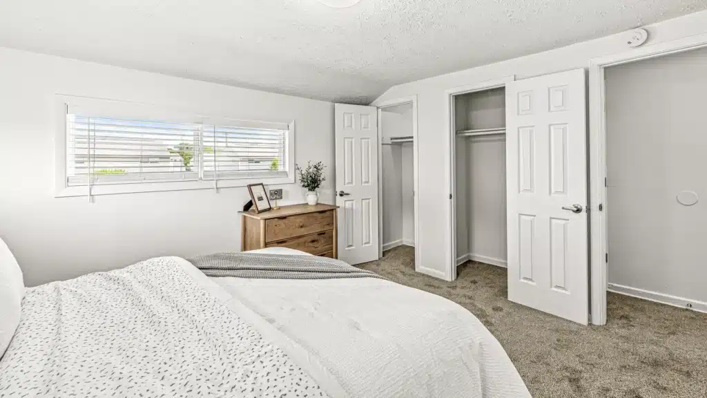A bright bedroom with a made bed, a wooden dresser with decor, open double closets, carpeted floor, and a window with blinds.