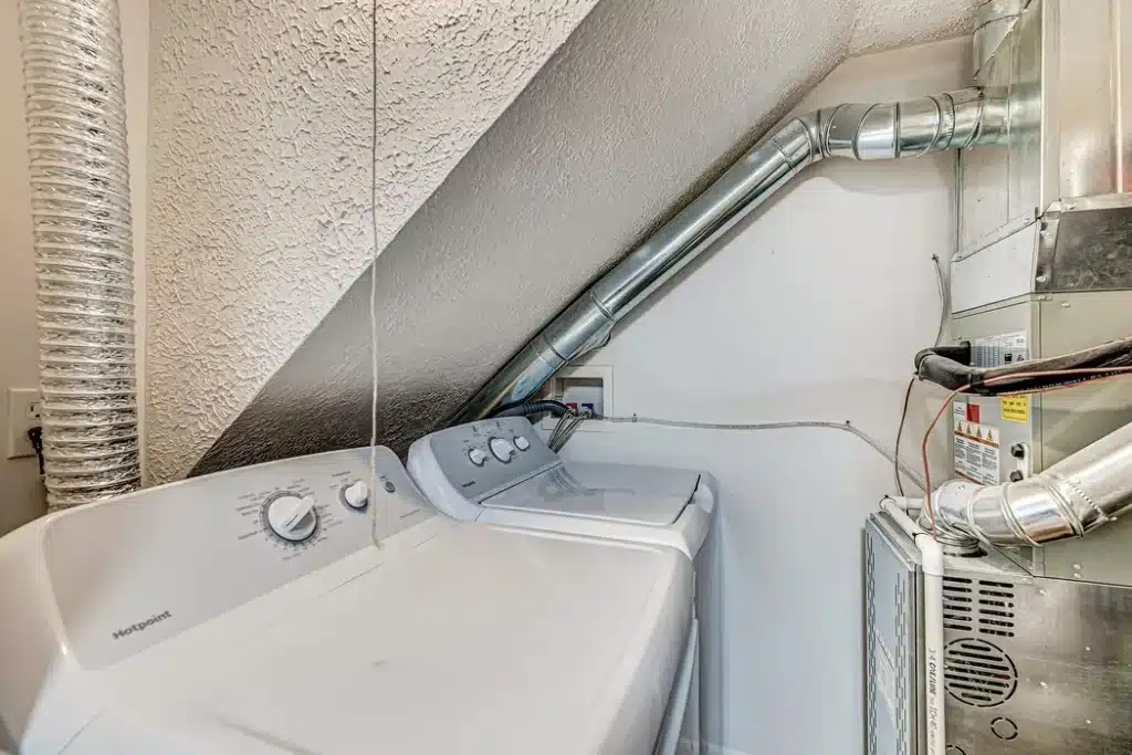 A washing machine and dryer sit side by side under a sloped ceiling next to an HVAC unit with exposed ducts in a small laundry area.